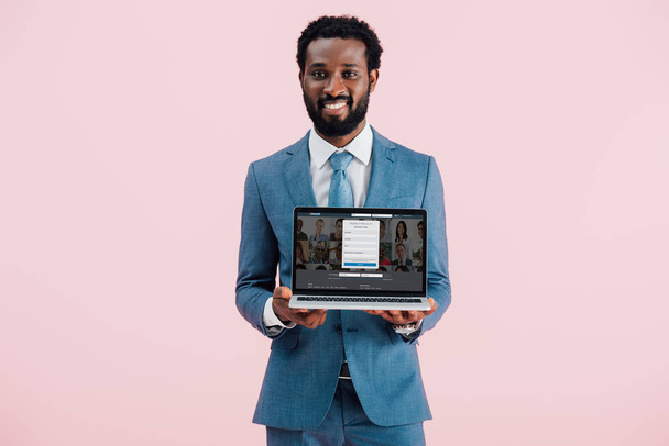 KYIV, UKRAINE - MAY 17, 2019: smiling african american businessman showing laptop with linkedin website, isolated on pink - Foto, immagini