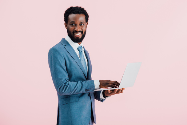 χαρούμενος Αφρικανός Αμερικανός επιχειρηματίας χρησιμοποιώντας laptop απομονωθεί σε ροζ - Φωτογραφία, εικόνα