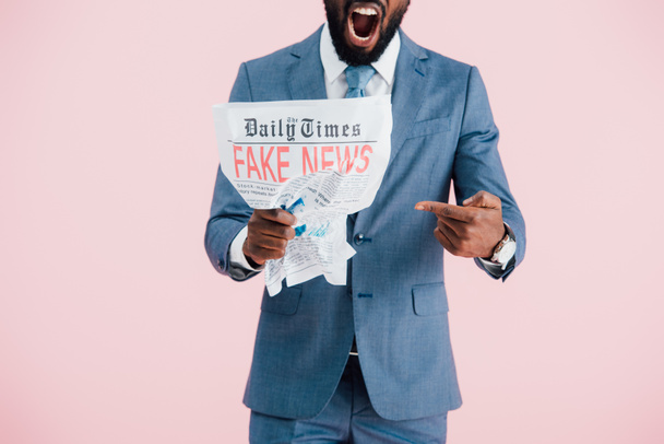ピンクに隔離された偽のニュースで新聞を保持する攻撃的なアフリカ系アメリカ人ビジネスマンのトリミングされたビュー - 写真・画像