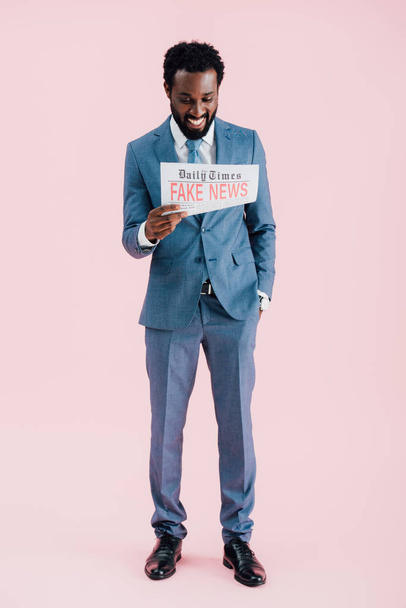 χαρούμενο αφρικανικό αμερικανικό επιχειρηματία ανάγνωση εφημερίδα με ψεύτικα νέα απομονώθηκαν σε ροζ - Φωτογραφία, εικόνα