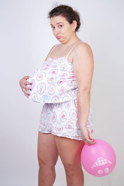 middelbare leeftijd meisje wil echt zwanger te raken. vrouw staande in pyjama met een lucht ballon onder een t-shirt op een witte achtergrond in de Studio - Foto, afbeelding