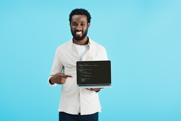 KYIV, UKRAINE - MAY 17, 2019: smiling african american man pointing at laptop with html code, isolated on blue - Photo, image