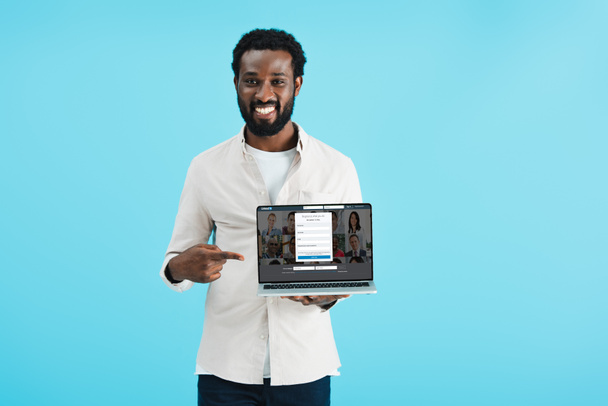 kyiv, ukraine - 17. Mai 2019: lächelnder afrikanisch-amerikanischer Mann zeigt auf Laptop mit linkedin Webseite, isoliert auf blau - Foto, Bild