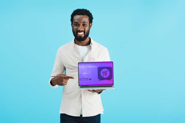 χαμογελαστός Αφρικανός Αμερικανός άνθρωπος δείχνοντας στο φορητό υπολογιστή με Web Shopping ιστοσελίδα απομονώνεται σε μπλε - Φωτογραφία, εικόνα