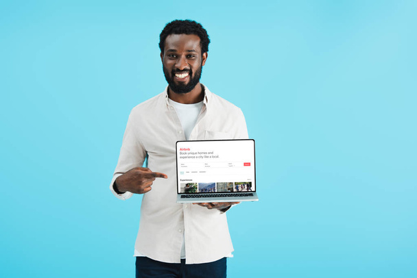 KYIV, UCRAINA - 17 MAGGIO 2019: uomo afroamericano sorridente che punta al laptop con sito airbnb, isolato sul blu
 - Foto, immagini