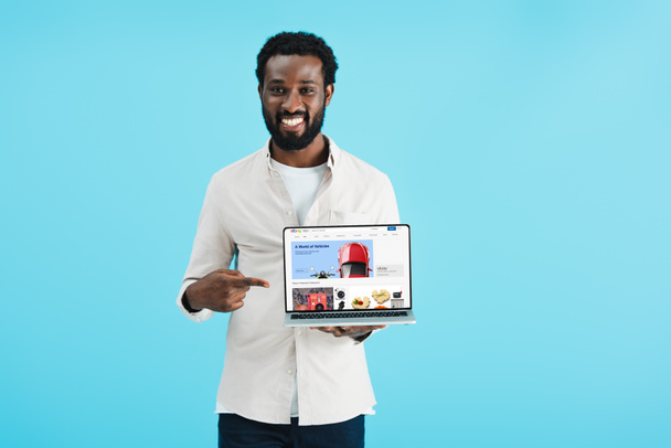 KYIV, UKRAINE - 17 MAI 2019 : homme afro-américain souriant pointant vers un ordinateur portable avec le site Web ebay, isolé sur bleu
 - Photo, image