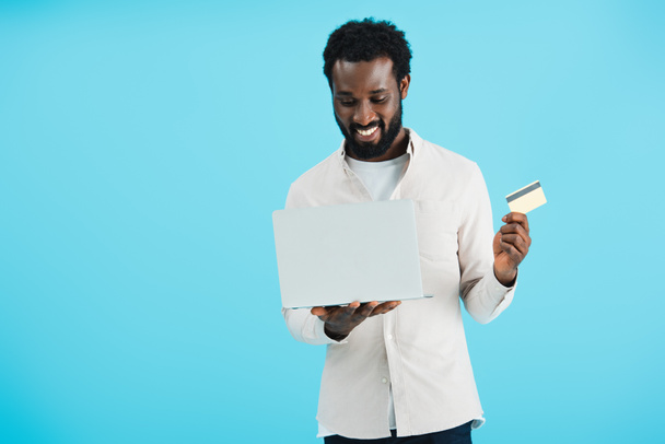 sonriente afroamericano hombre de compras en línea con tarjeta de crédito y portátil aislado en azul
 - Foto, Imagen