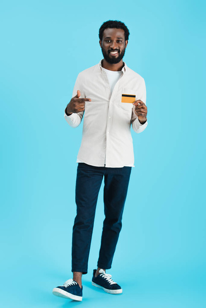 glimlachend Afro-Amerikaanse man wijst op creditcard geïsoleerd op blauw - Foto, afbeelding