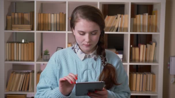 Closeup portrait of young pretty female student texting on the tablet looking at camera in the university library indoors - Materiaali, video