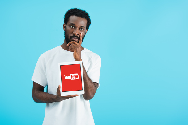 kyiv, ukraine - 17. Mai 2019: nachdenklicher afrikanisch-amerikanischer Mann zeigt digitales Tablet mit Youtube-App, isoliert auf blauem Grund - Foto, Bild