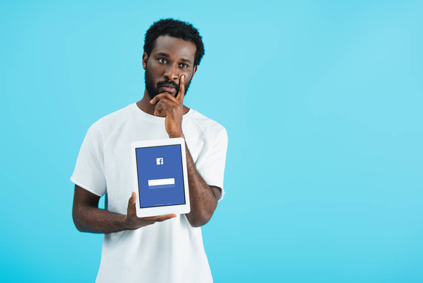 KYIV, UKRAINE - MAY 17, 2019: thoughtful african american man showing digital tablet with facebook app, isolated on blue - Foto, afbeelding