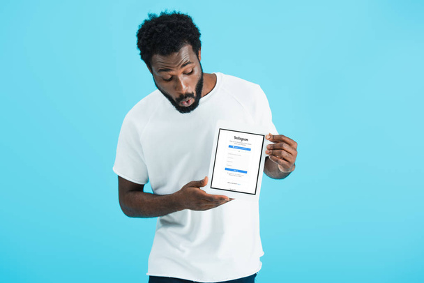 kyiv, ukraine - 17. Mai 2019: schockierter afrikanisch-amerikanischer Mann schaut auf digitales Tablet mit Instagram-App, isoliert auf blau - Foto, Bild