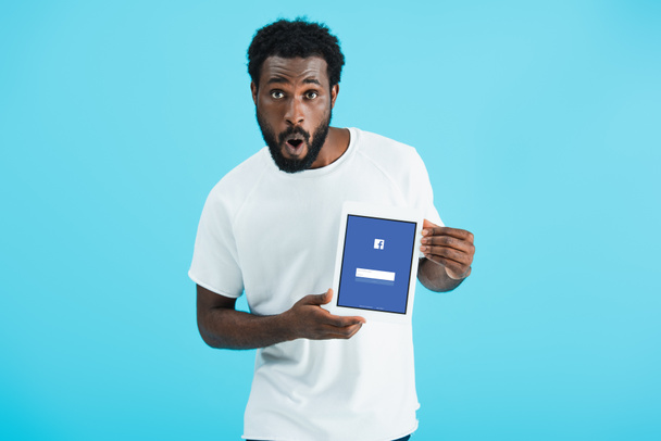 KYIV, UCRANIA - 17 de mayo de 2019: sorprendido hombre afroamericano mostrando tableta digital con aplicación de facebook, aislado en azul
 - Foto, Imagen
