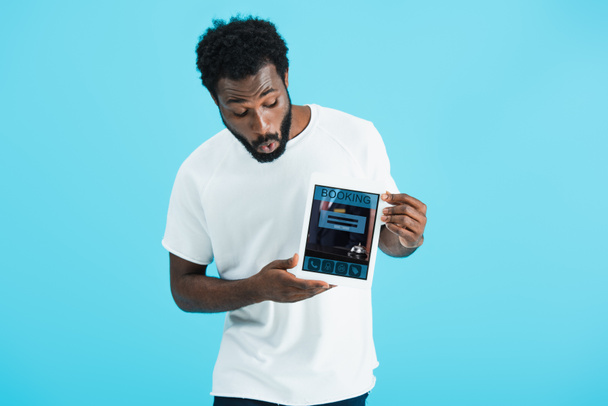 wstrząśnięty African American Man pokazując cyfrowy tablet z aplikacji rezerwacji, izolowane na niebiesko - Zdjęcie, obraz