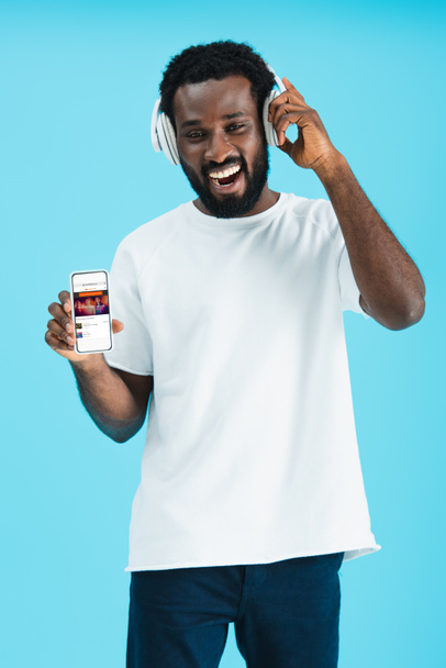 KYIV, UKRAINE - MAY 17, 2019: smiling african american man listening music with headphones and showing smartphone with soundcloud app, isolated on blue - 写真・画像