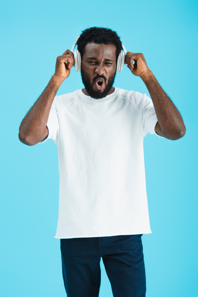 excited african american man singing and listening music with headphones, isolated on blue - Фото, изображение