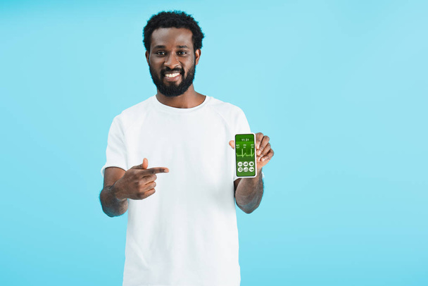 smiling african american man pointing at smartphone with health app, isolated on blue - Foto, imagen