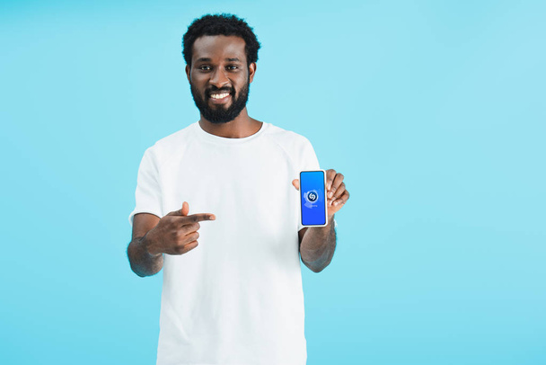KYIV, UKRAINE - MAY 17, 2019: smiling african american man pointing at smartphone with shazam app, isolated on blue - Fotografie, Obrázek