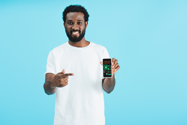 lächelnder Afrikaner, der auf ein Smartphone mit Marketing-Analyse zeigt, isoliert auf blauem Grund - Foto, Bild