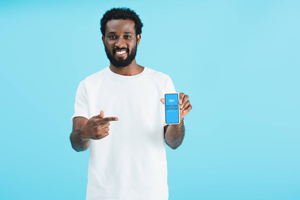 KYIV, UKRAINE - MAY 17, 2019: smiling african american man pointing at smartphone with skype app, isolated on blue - Fotoğraf, Görsel