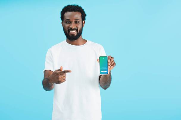 KYIV, UKRAINE - MAY 17, 2019: smiling african american man pointing at smartphone with twitter app, isolated on blue - Foto, imagen