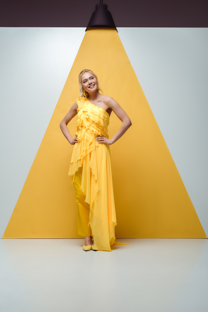 cheerful blonde girl smiling while standing with hands on hips on white and yellow  - Fotó, kép