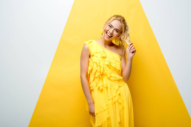 happy blonde girl touching hair while posing on white and yellow  - Foto, afbeelding