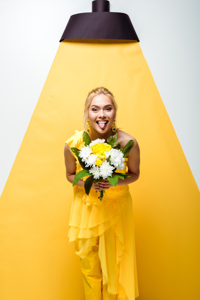 happy young woman showing tongue and holding bouquet of flowers on white and yellow  - Foto, immagini
