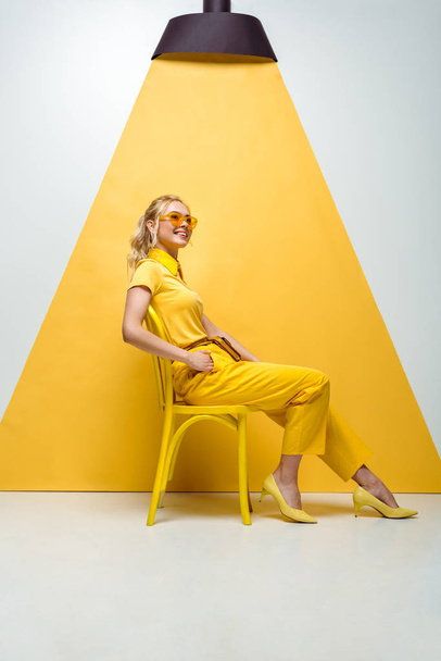 happy blonde woman in sunglasses sitting on chair on white and yellow  - Фото, зображення