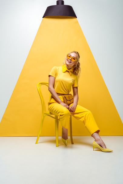 positive blonde woman in sunglasses sitting on chair on white and yellow  - Fotó, kép