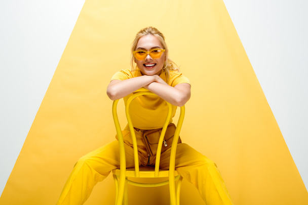 cheerful blonde woman in sunglasses sitting on chair on white and yellow  - Valokuva, kuva