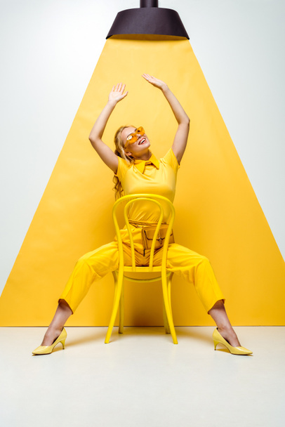 happy blonde woman in sunglasses sitting on chair with hands above head and looking at lamp on yellow and white - Foto, imagen