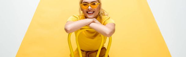 panoramic shot of happy blonde woman in sunglasses sitting on chair on yellow and white - Foto, immagini