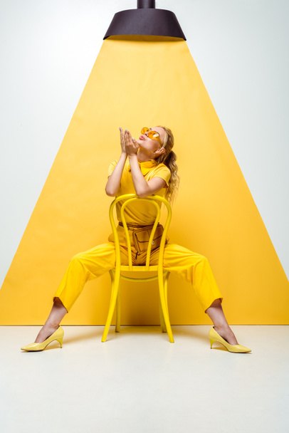 blonde woman in sunglasses sitting on chair and sending air kiss while looking at lamp on yellow and white - Zdjęcie, obraz
