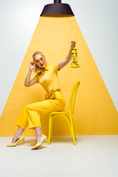 attraktive blonde Frau, die Sonnenbrille anfasst, während sie auf einem Stuhl sitzt und eine Vintage-Lampe in weiß und gelb hält  - Foto, Bild