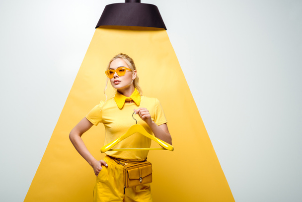ελκυστική γυναίκα στα γυαλιά ηλίου κρατώντας κρεμάστρα και στέκεται με το χέρι στην τσέπη σε λευκό και κίτρινο  - Φωτογραφία, εικόνα
