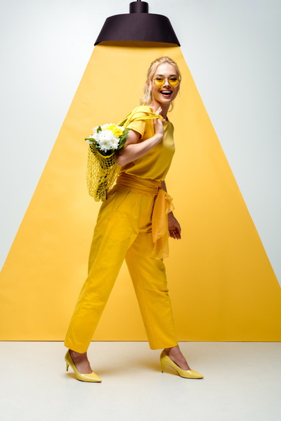 cheerful blonde woman holding string bag with flowers while posing on white and yellow  - Zdjęcie, obraz