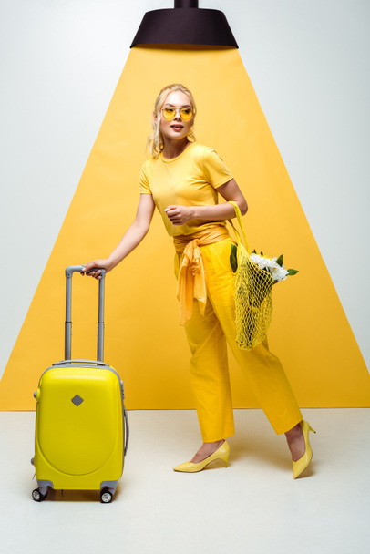 attractive blonde girl posing near luggage and holding reusable bag with flowers on white and yellow  - Φωτογραφία, εικόνα