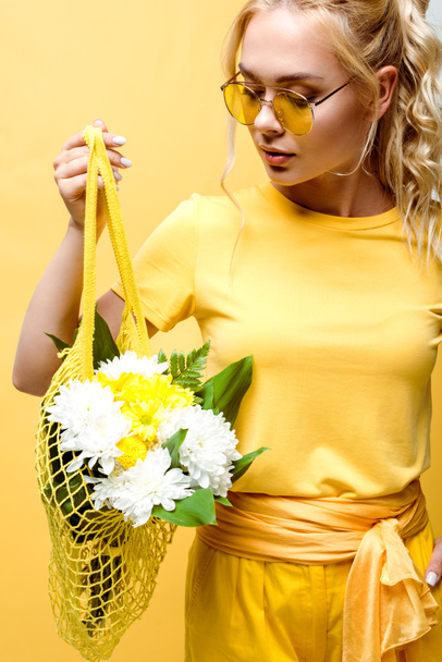 attractive young woman holding string bag with flowers isolated on yellow  - Fotó, kép