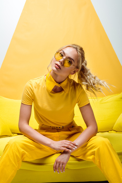 attractive blonde woman in sunglasses sitting on sofa and looking at camera on white and yellow  - Foto, immagini