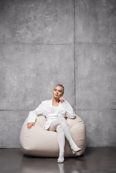 pensive blonde woman sitting on soft bean bag chair on grey  - Fotó, kép