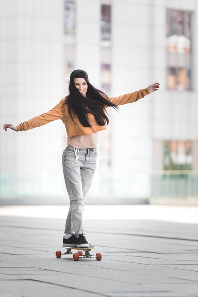 brunette woman with outstretched hands looking at camera, riding on skateboard in city - Foto, Imagem