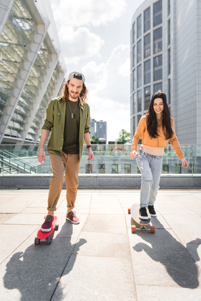 happy man looking at camera and riding on skateboards on roof with beautiful woman - Photo, Image