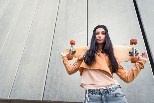 low angle view of brunette woman in casual wear standing near concentrate wall, holding skateboard behind back, looking at camera - Photo, Image