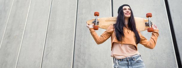 низкий угол обзора женщины в повседневной одежде, стоящей рядом со стеной концентрата со скейтбордом в руках, глядя в сторону
 - Фото, изображение