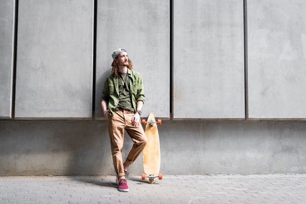 handsome man in casual wear standing near concentrate wall and skateboard, looking away - Foto, afbeelding