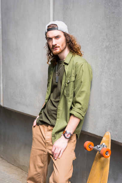 pohledný muž stojící blízko soustředného opevnění a skateboard, pohled na kameru - Fotografie, Obrázek