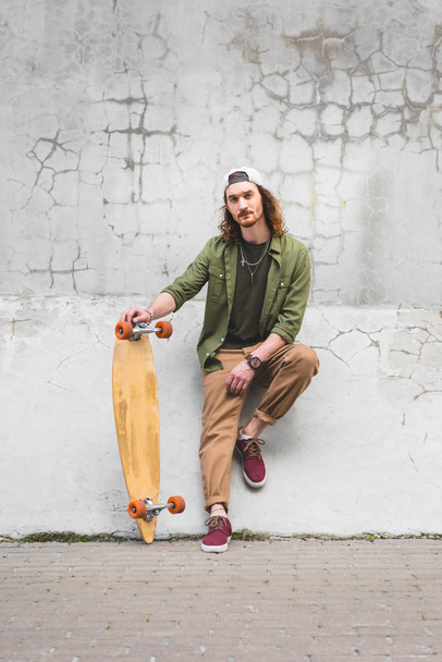 confident man in casual wear looking at camera, standing near wall with skateboard in hand - Fotó, kép