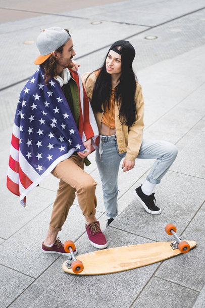 hoge hoek uitzicht van de mens met Amerikaanse vlag op schouders kijken naar brunette vrouw, staande in de buurt van skateboard - Foto, afbeelding
