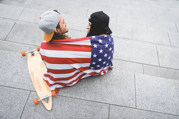 スケートボードの近くの階段に座って、お互いを見て、肩にアメリカの旗を持つカップルの高い角度のビュー - 写真・画像